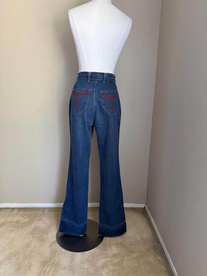 K&ouml;nnte beinhalten: Dunkelblaue Schlaghose aus Jeansstoff mit roten N&auml;hten auf den Ges&auml;&szlig;taschen. Die Jeans wird auf einer wei&szlig;en Schaufensterpuppe pr&auml;sentiert. Die Jeans hat eine hohe Taille und einen ausgestellten Bein-Design, typisch f&uuml;r die Mode der 1970er Jahre.