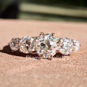 May include: A rose gold ring featuring five round-cut, clear gemstones. The central gemstone is larger than the others. The ring is set against a blurred background, with the text "DiamondHolicUS" visible.