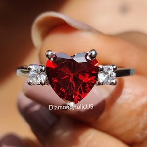 May include: A silver ring with a heart-shaped, deep red gemstone at its centre, with two smaller, square-cut white gemstones on either side. The ring is held up, highlighting its design and sparkle.