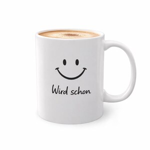 Könnte beinhalten: Weiße Keramik-Kaffeetasse mit einem schwarzen Smiley und dem deutschen Ausdruck "Wird schon" auf der Vorderseite. Die Tasse ist mit einem hellbraunen Getränk gefüllt, möglicherweise Kaffee oder Tee.