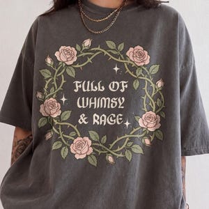 Op de afbeelding: Een antracietgrijs t-shirt met korte mouwen en een cirkelvormige krans van groene doornige ranken en lichtroze rozen. In het midden staat de tekst "FULL OF WHIMSY & RAGE" in een decoratief lettertype, geaccentueerd door kleine vierpuntige sterren.