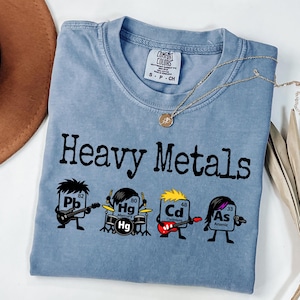 May include: A pale blue Comfort Colors t-shirt featuring the words "Heavy Metals" and cartoon illustrations of elements playing instruments. A gold necklace is draped across the shirt.