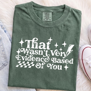 Op de afbeelding: Een bosgroen t-shirt met witte gezeefdrukte tekst die That Wasn't Very Evidence Based Of You luidt, vergezeld door grafische elementen waaronder een bliksemschicht, verschillende vierpuntige sterren en een dambordpatroon onderaan de tekst.