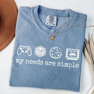 May include: A blue Comfort Colors t-shirt with the phrase "my needs are simple" and icons of a game controller, a 20-sided die, a cookie, and a video game cartridge. A gold necklace is draped across the shirt.