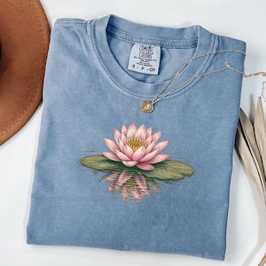 May include: A blue short-sleeved t-shirt featuring a graphic of a pink lotus flower resting on a green lily pad with a water reflection. The shirt has a Comfort Colors brand label at the neck with size S P CH printed on it and a gold-toned necklace.