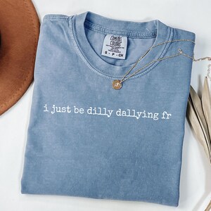 Könnte beinhalten: Ein blaues kurzärmeliges T-Shirt mit Rundhalsausschnitt und verwaschener Optik. Auf der Brust ist der weiße Schriftzug "i just be dilly dallying fr" aufgedruckt. Eine kleine runde Anhängerkette mit Rosenmotiv liegt auf dem Stoff des T-Shirts.
