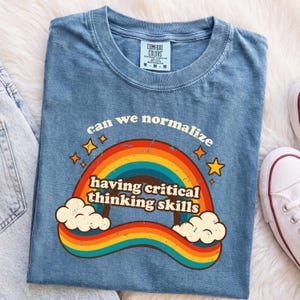 Könnte beinhalten: Ein blaues kurzärmeliges T-Shirt mit einer Grafik aus Regenbogen, Wolken und gelben Sternen. Der Text lautet "can we normalize having critical thinking skills". Das T-Shirt hat einen Rundhalsausschnitt und ein Comfort Colors Markenetikett.