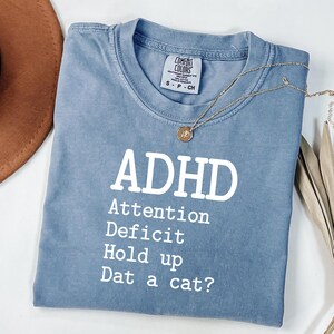 以下が含まれることがあります： 大きな白い文字で「ADHD」と書かれた青いComfort ColorsのTシャツ。その下に「Attention Deficit Hold up Dat a cat?」という言葉が印刷されています。ペンダント付きの金のネックレスがシャツの上にかけられています。