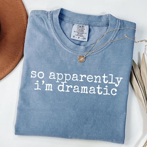 Könnte beinhalten: Ein blaues Comfort Colors T-Shirt mit dem weißen Schriftzug "so apparently i'm dramatic". Eine goldene Halskette mit Anhänger liegt über dem Shirt. Ein brauner Hut ist teilweise in der oberen linken Ecke zu sehen.