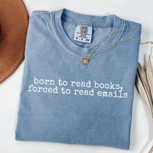 May include: A blue t-shirt with the text "born to read books, forced to read emails" in white. A gold necklace with a pendant is draped across the shirt. The shirt is folded on a white surface.