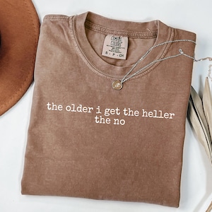 May include: A brown short-sleeved t-shirt with a crew neckline and a Comfort Colors brand label at the inner collar. The shirt features white printed text that reads: the older i get the heller the no. A thin silver-toned necklace rests on the fabric.