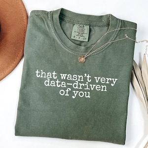 May include: An olive green short-sleeved t-shirt with a crew neckline and the white text that wasn't very data-driven of you printed on the front. The shirt features a Comfort Colors brand label at the inner neck and a thin gold-toned necklace.