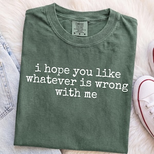 Pode incluir: Uma t-shirt de manga curta verde-floresta com decote redondo e o texto impresso em branco "i hope you like whatever is wrong with me" no peito. A t-shirt apresenta uma etiqueta da marca Comfort Colors no interior da gola.