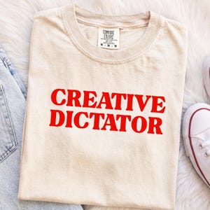Może przedstawiać: Beżowy t-shirt z krótkim rękawem i okrągłym dekoltem, z napisem "CREATIVE DICTATOR" wydrukowanym pogrubionymi czerwonymi wielkimi literami na klatce piersiowej. Koszulka posiada metkę marki Comfort Colors wewnątrz kołnierza wskazującą rozmiar M.