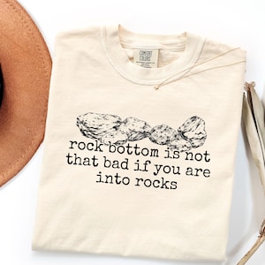 May include: A cream-colored short-sleeve t-shirt featuring a black ink illustration of several rocks above the text "rock bottom is not that bad if you are into rocks" printed in a typewriter-style font. The shirt has a Comfort Colors brand label.