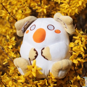 Puede incluir: Un juguete de peluche blanco con un cuerpo redondo, ojos marrones y una pequeña nariz naranja. El juguete está sentado en una cama de flores amarillas.