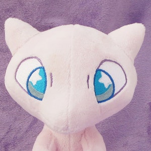 May include: Pink plush toy of a Pokemon character with large blue eyes and a white star shape in each eye. The toy has a soft, fuzzy texture.