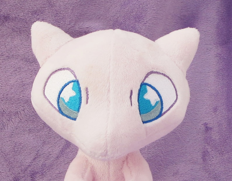 May include: Pink plush toy of a Pokemon character with large blue eyes and a white star shape in each eye. The toy has a soft, fuzzy texture.