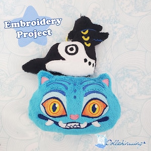 May include: Two plush toys: a blue cat-like creature with orange eyes and a black and white creature. The blue cat has a wide grin and pink accents. The black and white creature has yellow accents. The text "Embroidery Project" is in a blue star.