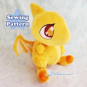 Könnte beinhalten: Ein gelber Plüschdrache mit orangenen Füßen und Flügeln. Der Drache hat große, schwarze Augen mit roten und weißen Details. Der Text "Sewing Pattern" ist in der oberen linken Ecke des Bildes sichtbar.