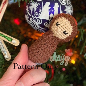May include: A brown crocheted baby Jesus ornament with a white face and black eyes. The ornament is hanging on a Christmas tree with a blue and white ornament behind it. The text "Pattern Only" is visible in the image.
