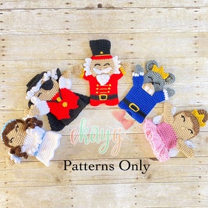 May include: Five crocheted finger puppets depicting characters from the Nutcracker ballet. The puppets are a Nutcracker soldier, a Mouse King, a Sugar Plum Fairy, a Clara, and a Pirate. The puppets are arranged in a circle on a wooden background. The text "Patterns Only" is visible below the puppets.