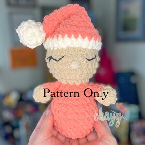 May include: A crocheted amigurumi doll wearing a pink and white hat. The doll is wearing a pink dress and has a beige face with closed eyes. The text "Pattern Only" is visible on the doll's dress.