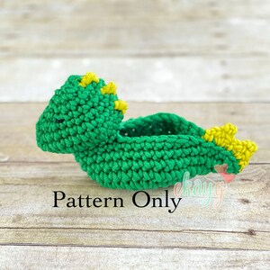 May include: Green crocheted dinosaur toy with yellow accents. The dinosaur is lying on its side with its head turned to the left. The text "Pattern Only" is visible below the dinosaur.