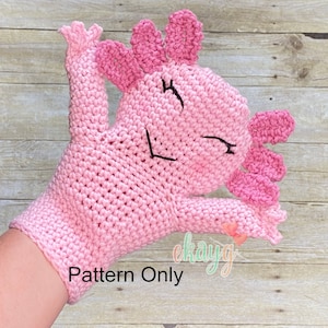 Può includere: Burattino di axolotl rosa all'uncinetto con occhi neri e una bocca sorridente. Il burattino è progettato per adattarsi a una mano e ha una coda lunga e due piccole braccia. Il testo "Pattern Only" è visibile nell'immagine.