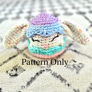 May include: A crocheted amigurumi toy in the shape of a bunny wearing a purple and blue hat. The bunny has a white body with multi-colored yarn accents. The toy is lying on a white and gray patterned surface. Pattern Only.