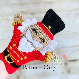 Peut inclure: Marionnette en crochet représentant un Casse-Noisette avec un uniforme rouge et or, une barbe blanche et un chapeau noir. La marionnette a les bras tendus, comme pour saluer. Le texte "Pattern Only" est visible sur l'image.