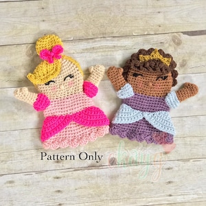 May include: Two crocheted princess dolls with different skin tones. The doll on the left has blonde hair and a pink dress. The doll on the right has brown hair and a purple dress. Both dolls have a crown on their head. The dolls are on a wooden background. Pattern Only.
