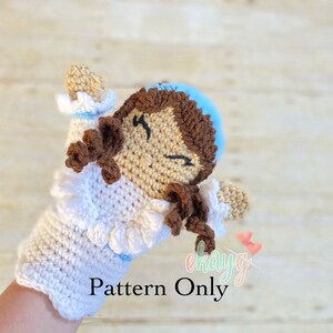 May include: Crochet doll puppet pattern. The doll has brown hair, a blue hat, and a white dress with a ruffle trim. The doll is attached to a white crochet hand puppet.