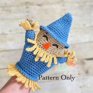 Può includere: Un burattino a forma di spaventapasseri all'uncinetto blu e beige con un volto sorridente, un cappello blu e frange gialle. Il testo "Pattern Only" è visibile.