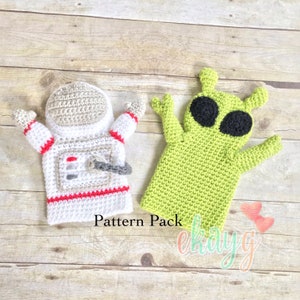 Op de afbeelding: Een haakpatroonpakket voor het maken van twee handpoppen: een astronaut en een alien. De astronautpop is wit met rode strepen en een grijs gezichtsmasker. De alienpop is groen met zwarte ogen.