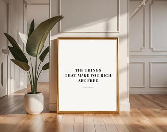 Może przedstawiać: Oprawiony wydruk z napisem "THE THINGS THAT MAKE YOU RICH ARE FREE" czcionką w kolorze czarnym. Rama jest złota, a wydruk jest eksponowany w pomieszczeniu z dużą rośliną w białej donicy i drewnianymi podłogami.