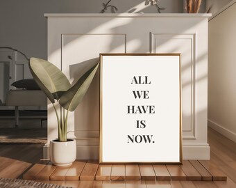 Könnte beinhalten: Ein gerahmter Druck mit dem Text "ALL WE HAVE IS NOW" in einem goldenen Rahmen. Der Druck steht auf einer Holzplattform, mit einer Topfpflanze links. Der Raum hat eine neutrale Farbpalette mit natürlichem Licht.