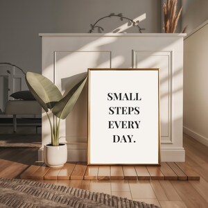 May include: A framed print with the text "SMALL STEPS EVERY DAY" in a gold frame. The print is displayed on a wooden platform, with a potted plant to the left. The background features a white wall and natural light.