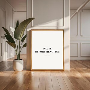 Puede incluir: Una impresión enmarcada con el texto "PAUSE BEFORE REACTING" en un marco dorado. La impresión está apoyada contra una pared blanca en una habitación con suelo de madera. Una gran planta en maceta está a la izquierda de la impresión.