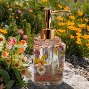 Könnte beinhalten: Ein klarer, rechteckiger Seifenspender mit goldfarbener Pumpe. Der Spender ist mit getrockneten Blumen und Blütenblättern gefüllt. Der Hintergrund zeigt einen lebendigen Garten mit bunten Blumen.