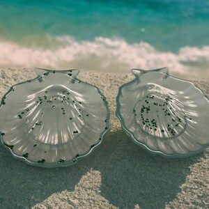 Könnte beinhalten: Zwei durchsichtige, muschelförmige Glasschalen mit strukturierter Oberfläche und dunkelgrünen Akzenten. Die Schalen liegen auf einem Sandstrand mit dem Ozean im Hintergrund. Die Schalen sind dekorativ.