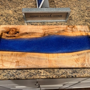 May include: A wooden serving tray with a vibrant blue resin river design. The tray has a rectangular shape with black handles and the text "HOME sweet HOME" in the background. The wood has a natural grain pattern.