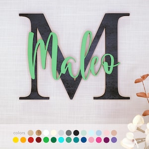 Könnte beinhalten: Wanddekoration aus Holz mit einem großen schwarzen Buchstaben "M" und dem Namen "Maleo" in grüner Schrift. Das Stück ist für die Wandmontage konzipiert, mit einer Auswahl an Farboptionen unten.