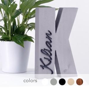 Op de afbeelding: Een grijze betonnen letter "K" met de naam "Kilian" in zwart schrift. De letter staat naast een witte potplant. De afbeelding toont ook een kleurenpalet met wit, grijs, zwart, beige en bruin.