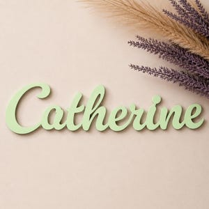Può includere: Un'insegna in legno tagliato color verde chiaro che riporta il nome Catherine in un carattere corsivo. Le lettere sono collegate in un unico pezzo, mostrando una finitura opaca liscia e un colore verde pastello uniforme su tutta la parola.