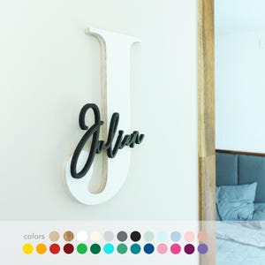 Könnte beinhalten: Ein weißer Holzbuchstabe "J" mit dem Namen "Julian" in schwarzer Schrift. Das Schild ist an einer weißen Wand angebracht, mit einem Blick auf ein Schlafzimmer im Hintergrund. Eine Farbpalette ist unten abgebildet.