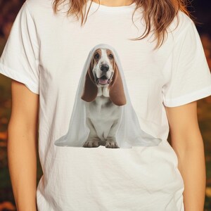 Könnte beinhalten: Weißes T-Shirt mit einem Basset Hound als Geist verkleidet. Der Hund hat lange braune Ohren und ist mit einem weißen Laken bedeckt. Der Hintergrund zeigt eine Frau, Kürbisse und Lichterketten, was auf ein Halloween-Thema hindeutet.