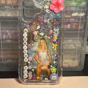 Può includere: Custodia trasparente per telefono con interno glitterato, adornata con vari ciondoli, tra cui un fiore rosa, stelle e una foto di una donna. Il testo "MIDNIGHT SUN" è scritto verticalmente sul lato.