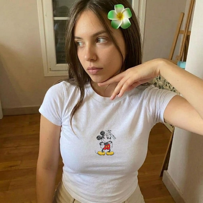 Puede incluir: Camiseta blanca de manga corta con un peque&ntilde;o gr&aacute;fico de Mickey Mouse en la parte delantera. El personaje de dibujos animados est&aacute; representado en negro, rojo, amarillo y blanco. Se ve un accesorio para el pelo de flores verdes y blancas.