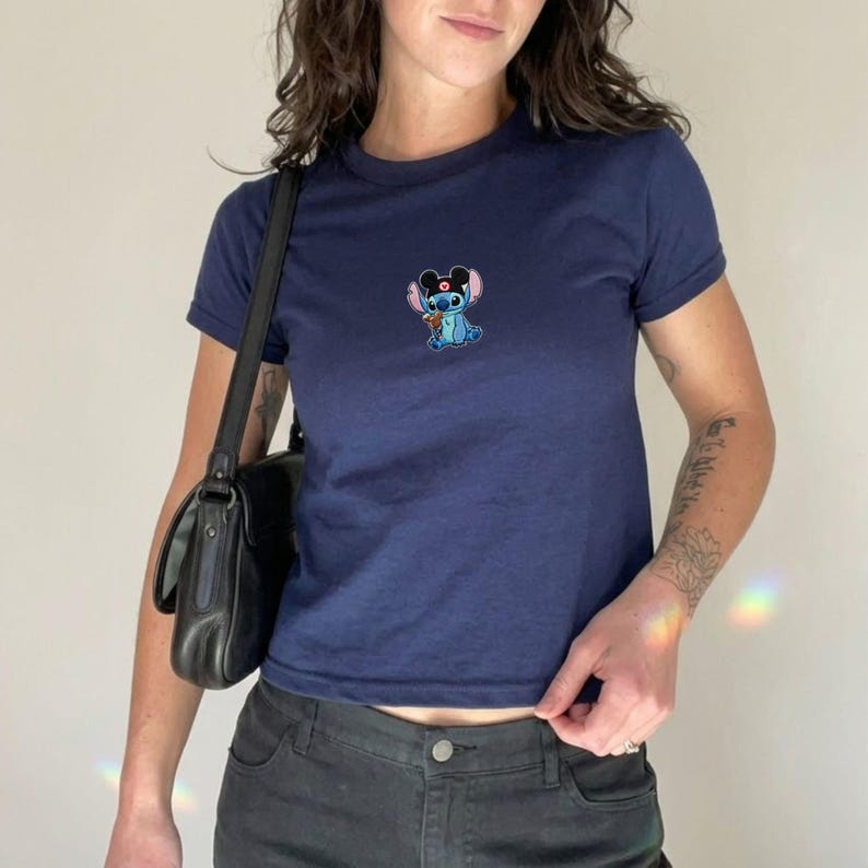 May include: Navy blue t-shirt with a small embroidered image of a blue cartoon character wearing a flower. The shirt has short sleeves and a crew neck. The shirt is paired with black jeans and a black shoulder bag.